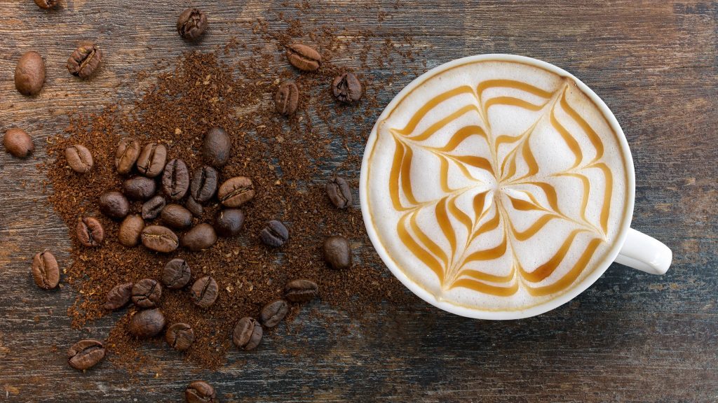 Aerial view of a coffee topped with foam with an intricate design. There are whole coffee and ground coffee mixed in an aesthetic manner to the left of the coffee cup.